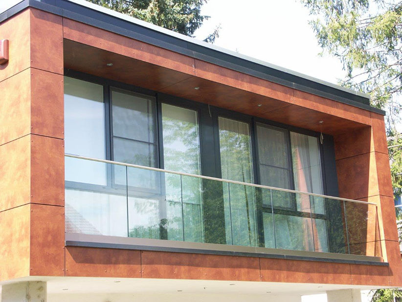 Das Foto zeigt ein Ganzglas-Balkongeländer mit Handlaufprofil. Es kommt bei einem Haus mit orange-/terrakottafarbener Fassade zum Einsatz.