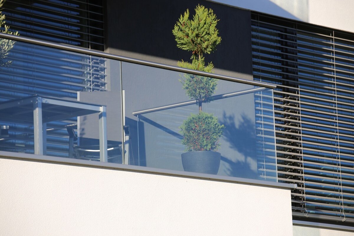 Vollglasgeländer / Ganzglasgeländer am Balkon eines modernen Hauses mit weißer Fassade und dunklen Jalousien